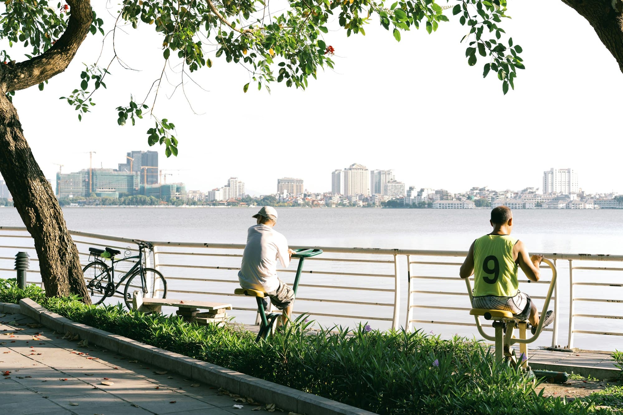 Two people exercising on outdoor fitness equipment beside a lake with a city skyline
