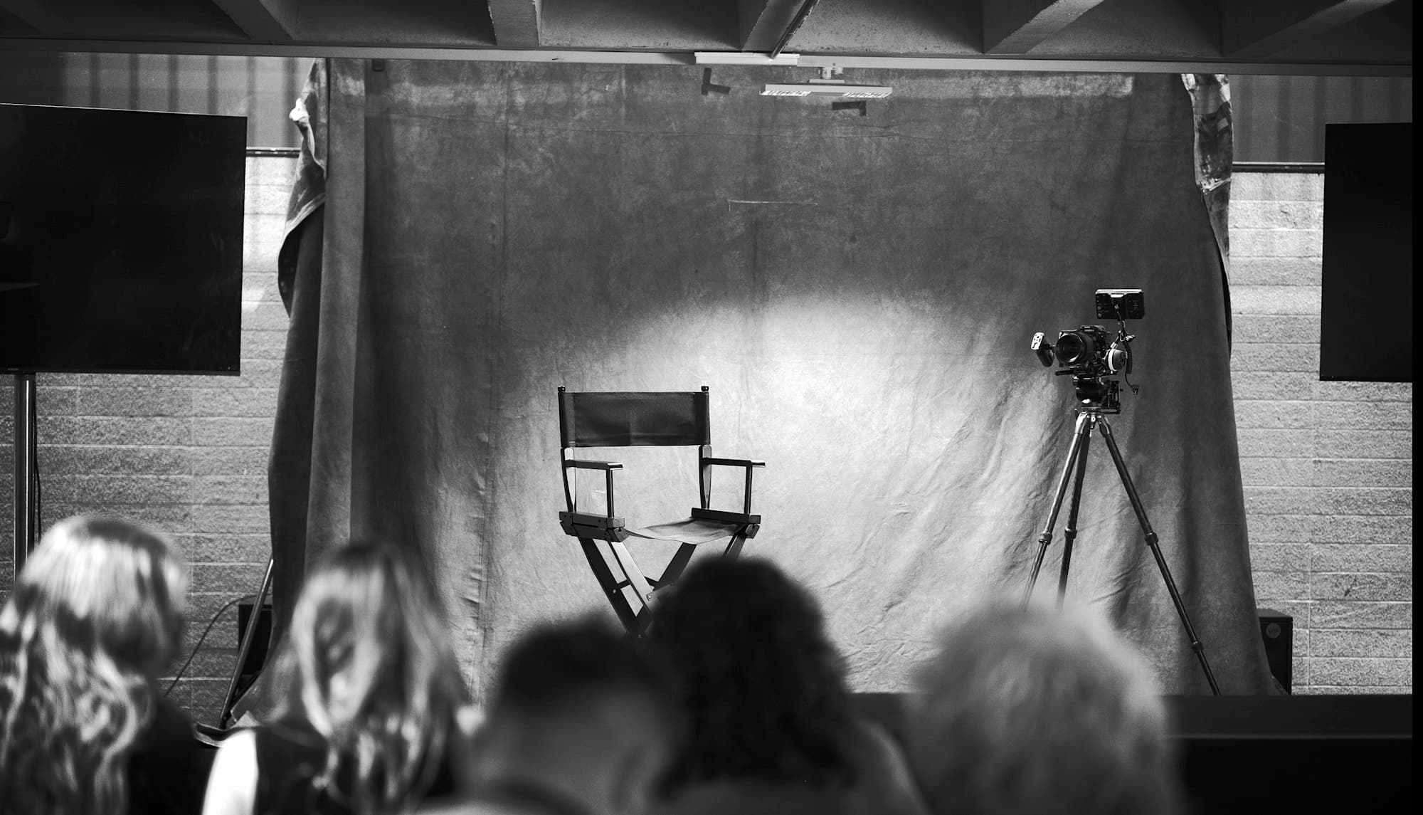 A director's chair and camera on a tripod on a lit stage facing an audience