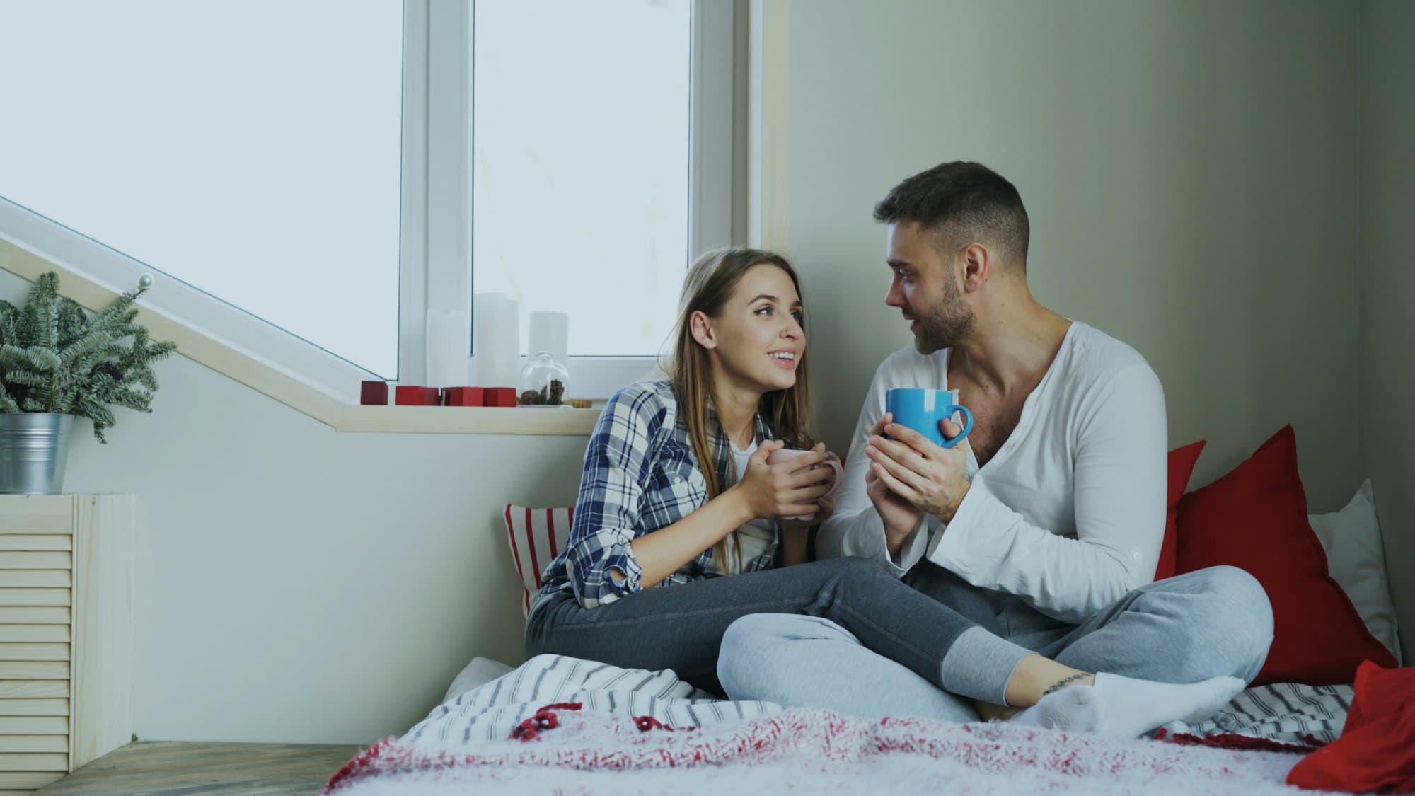 A couple sit close by a window holding mugs and talking warmly