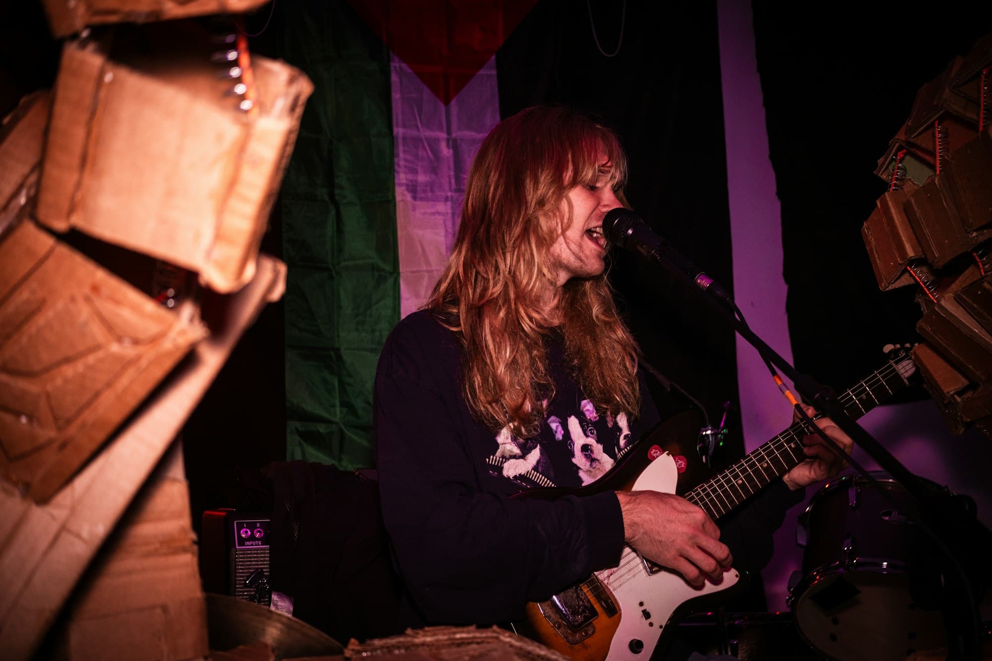 A long-haired musician singing and playing electric guitar on a small stage with cardboard sculptures