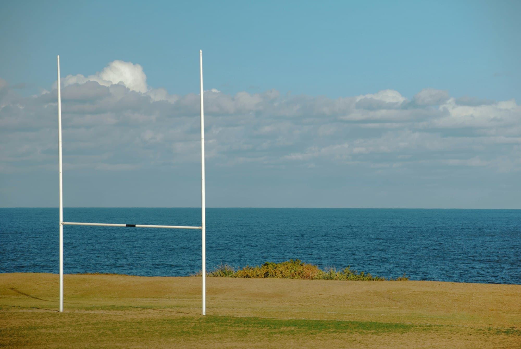 White rugby goal posts on a grassy field above a calm blue sea