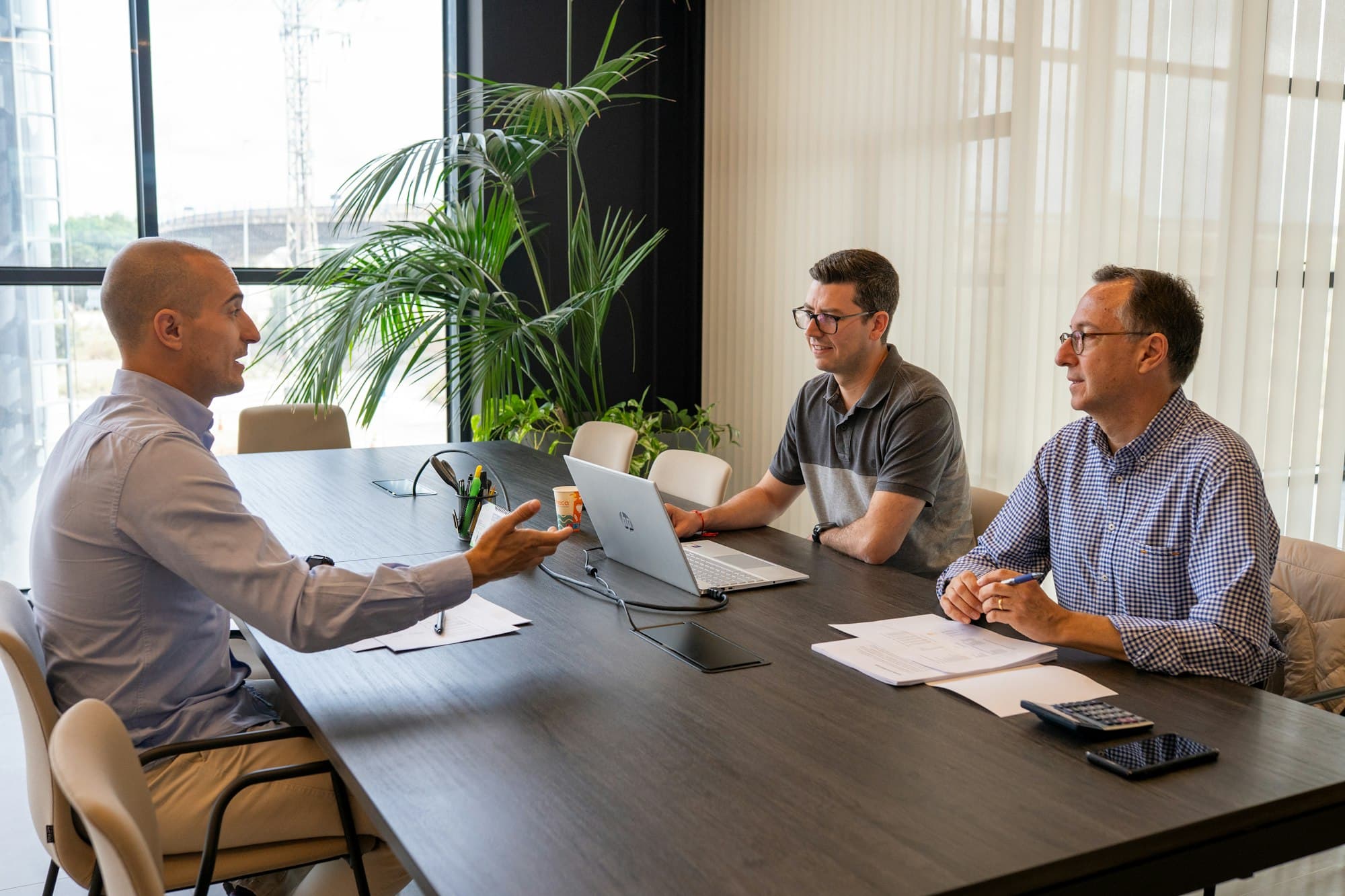 Three men in discussion around a dark wooden table in a bright office