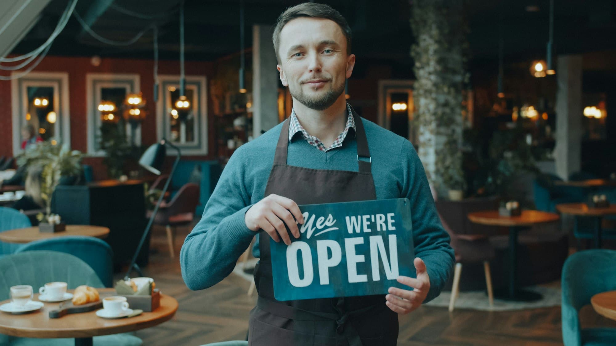 A cafe owner in an apron holding a sign that says Yes, we're open