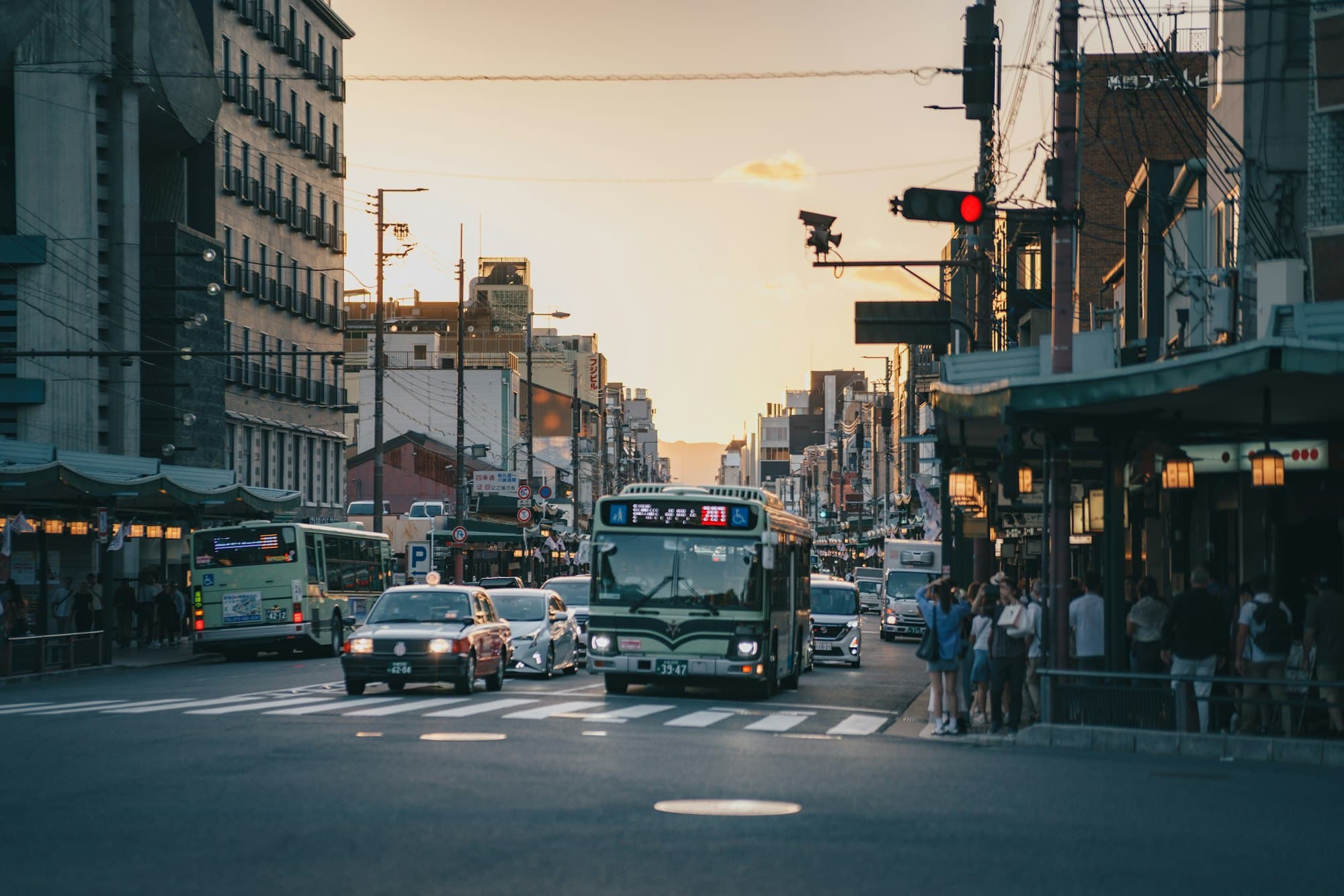 A city street at golden hour with a bus, cars and pedestrians waiting at a crossing
