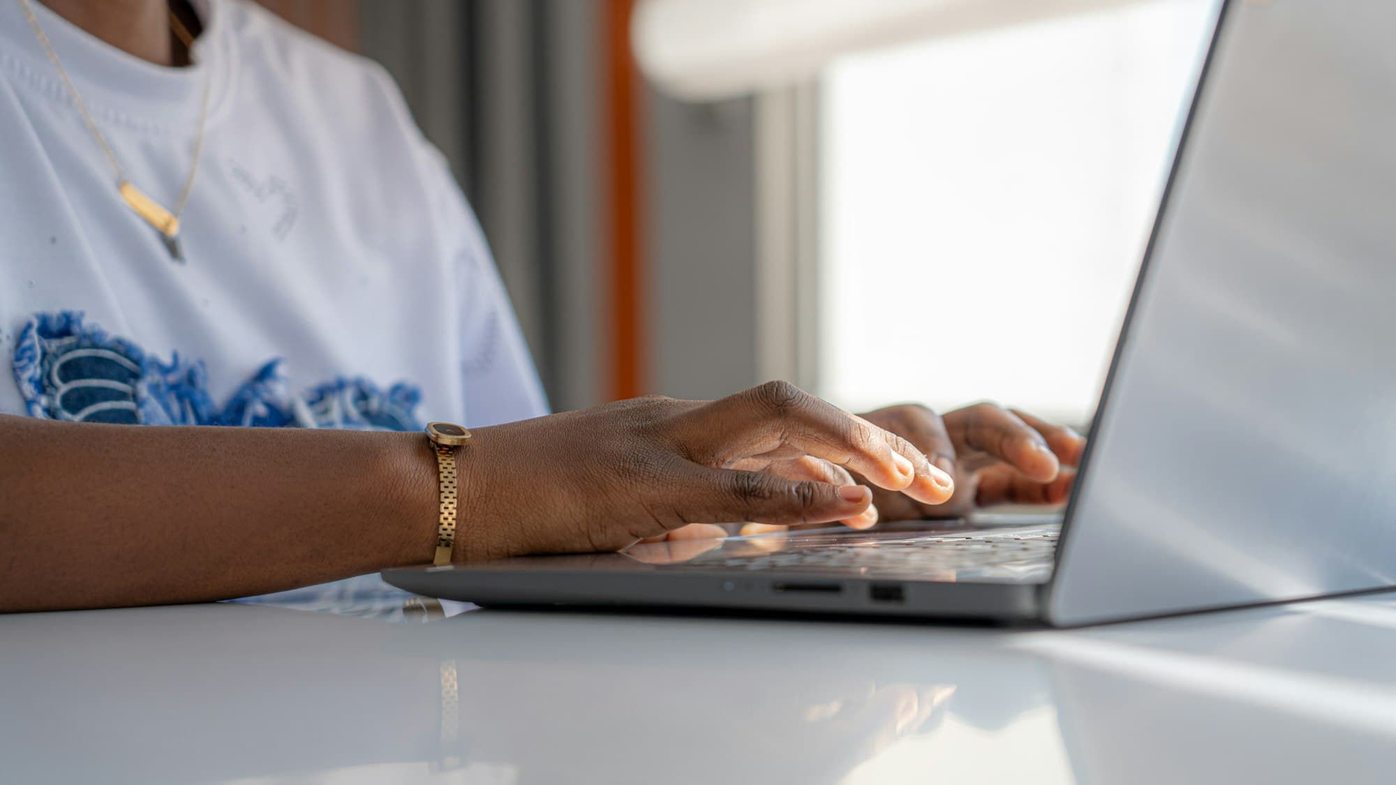 Hands typing on a laptop at a white desk in a sunlit room