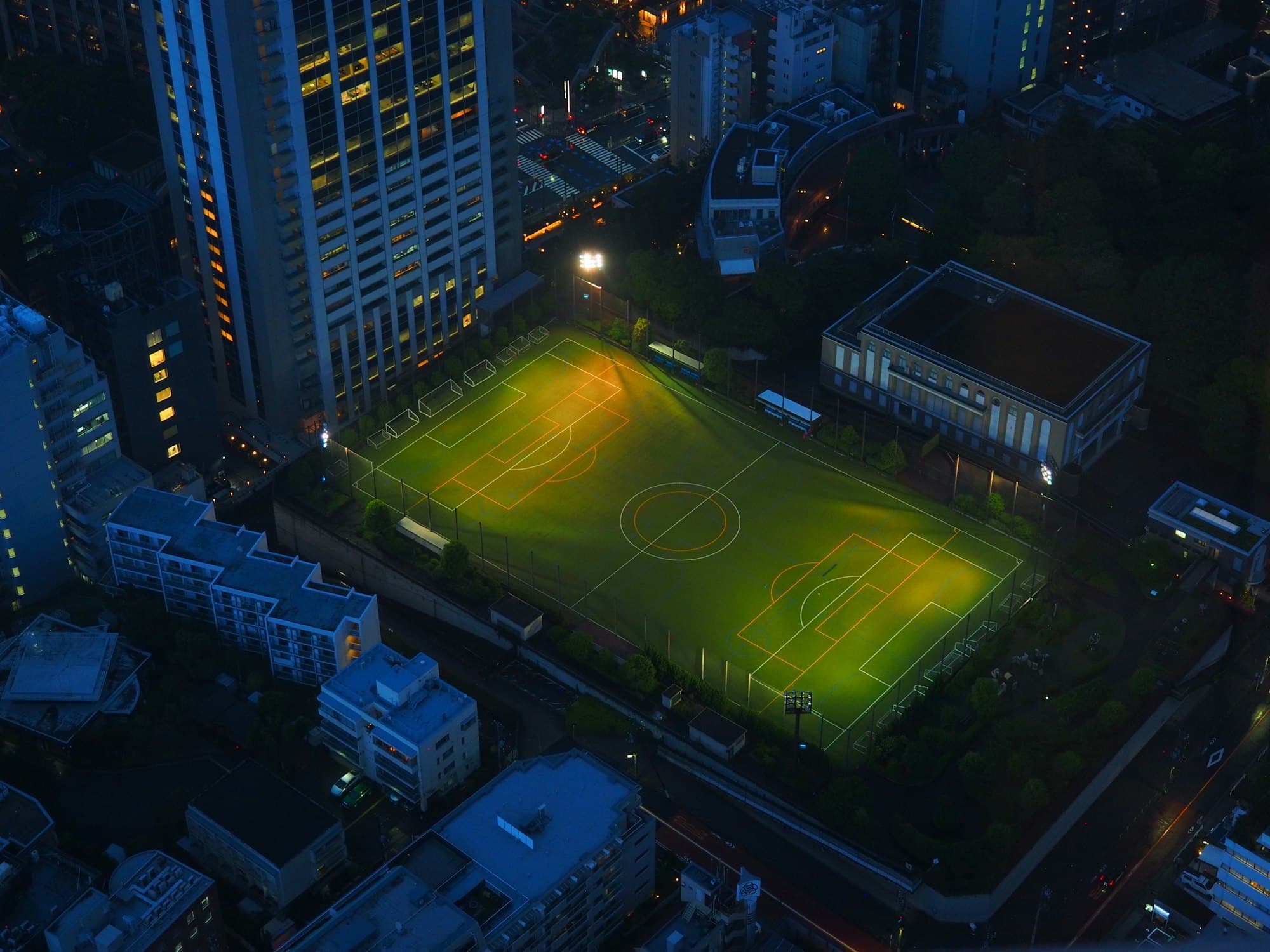 An aerial view of a brightly lit soccer field surrounded by city buildings at night