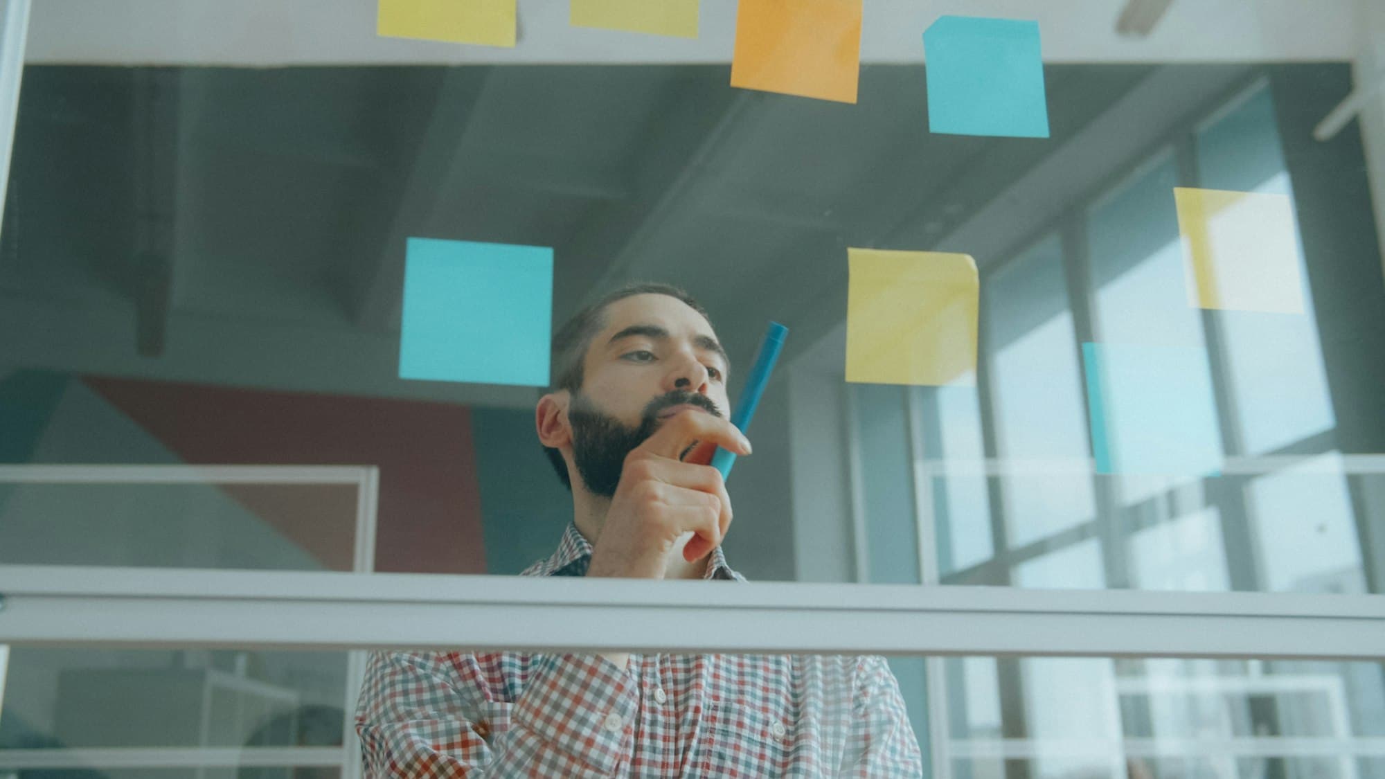 A man with a marker thinks behind a glass wall covered in blank sticky notes