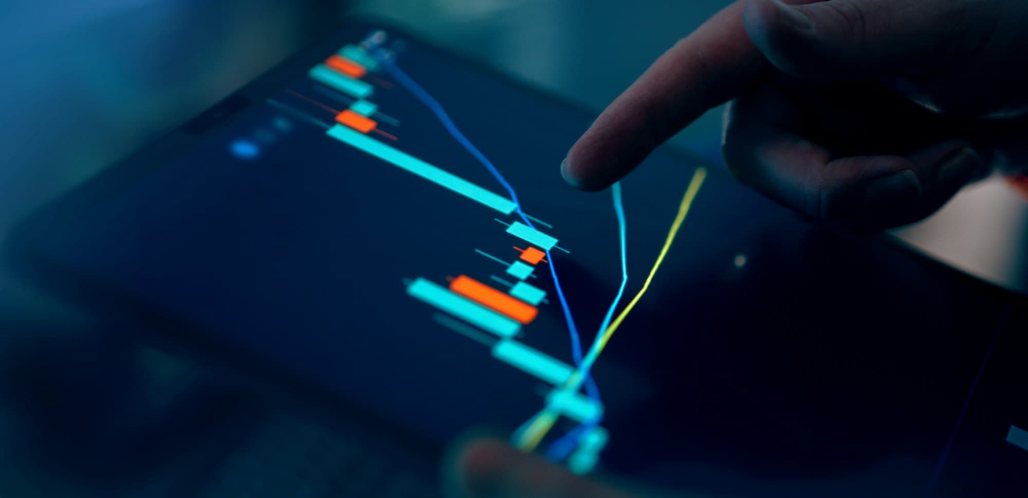 A finger pointing at candlestick charts on a dark tablet screen
