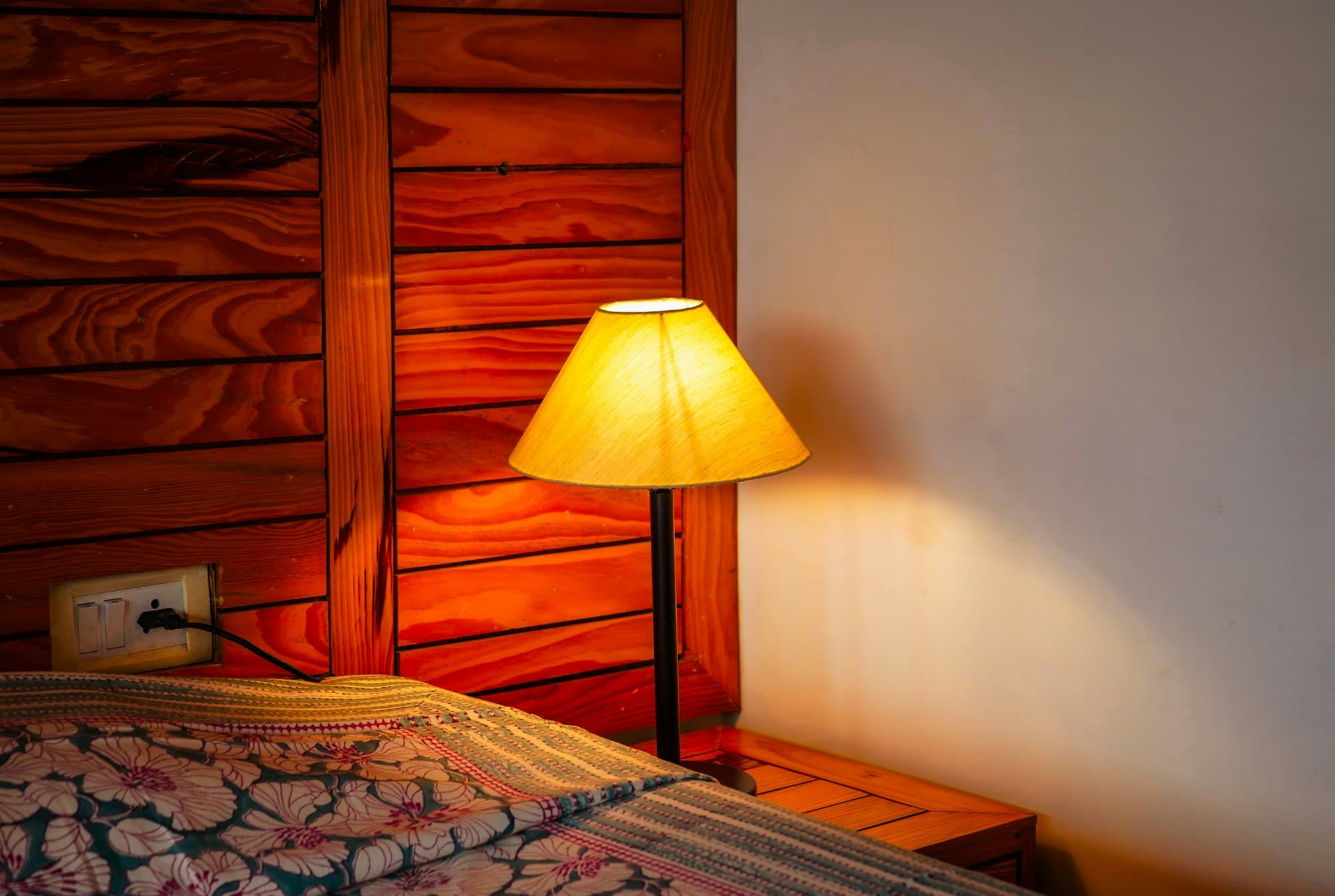 A lit table lamp with a yellow shade beside a bed with floral bedding