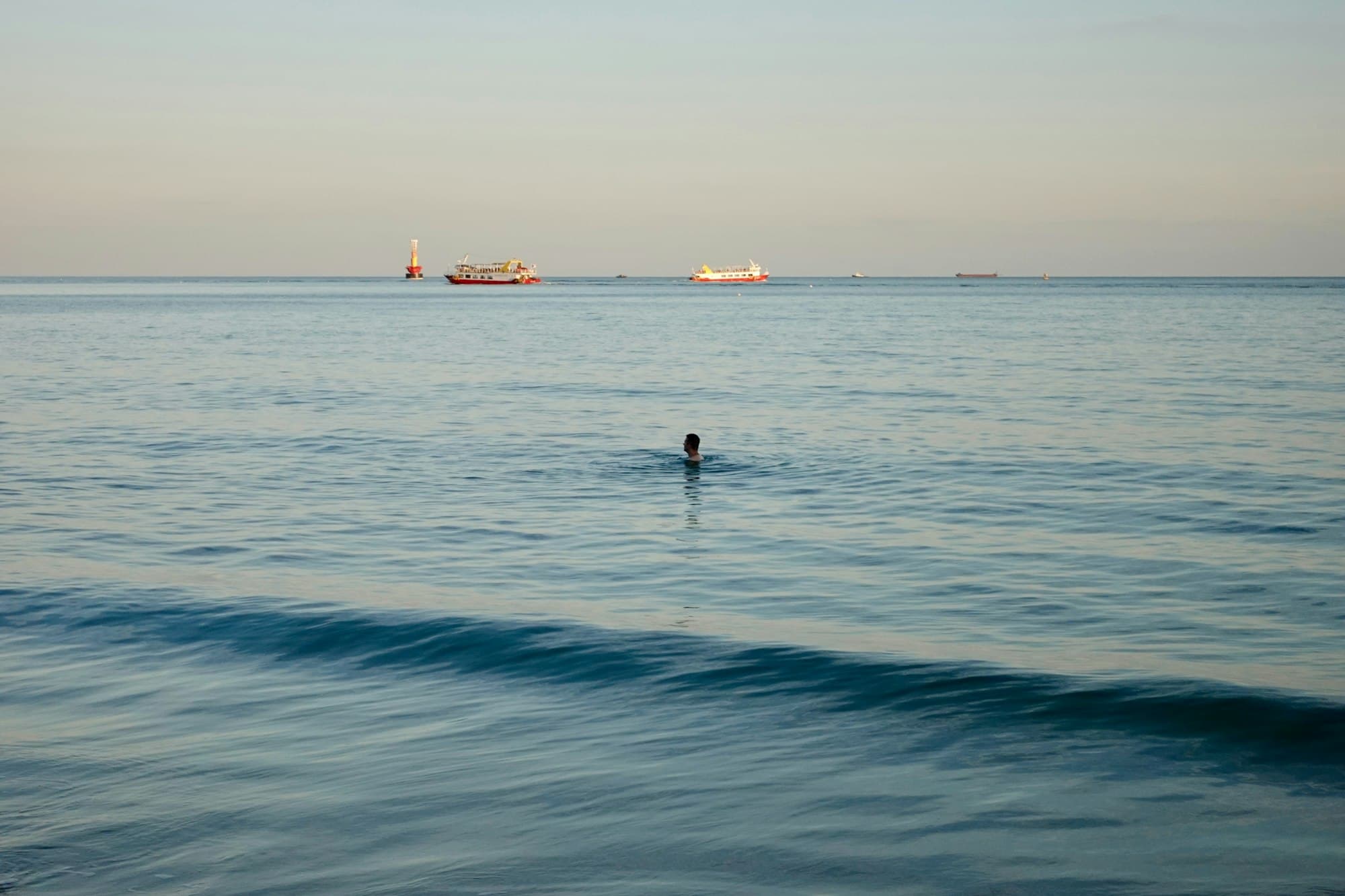 A lone swimmer in calm open water with boats on the horizon