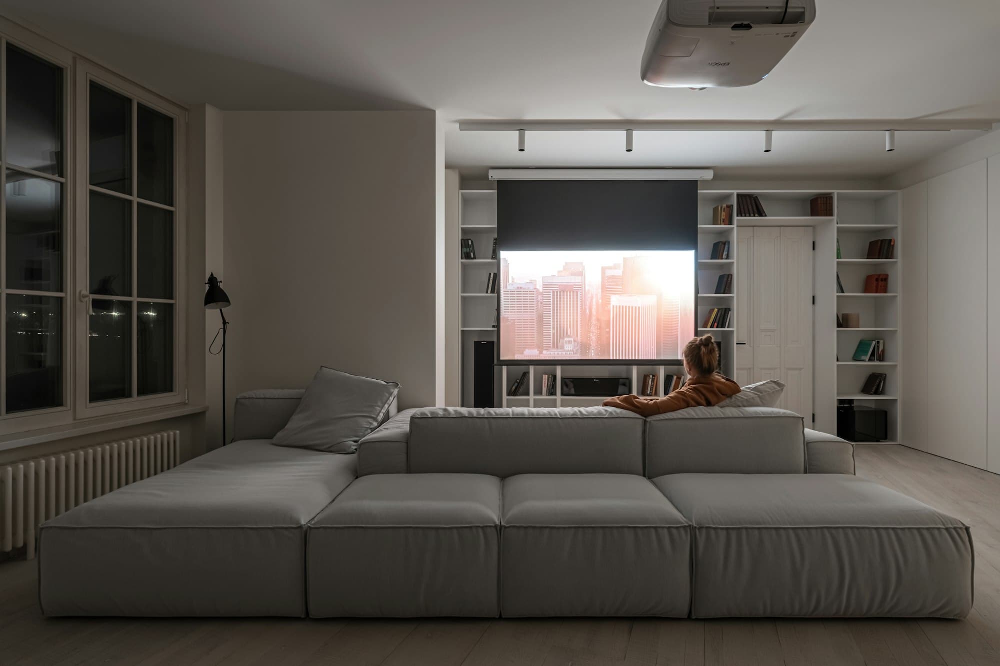 A person on a large grey sofa watching a projection screen in a dim living room