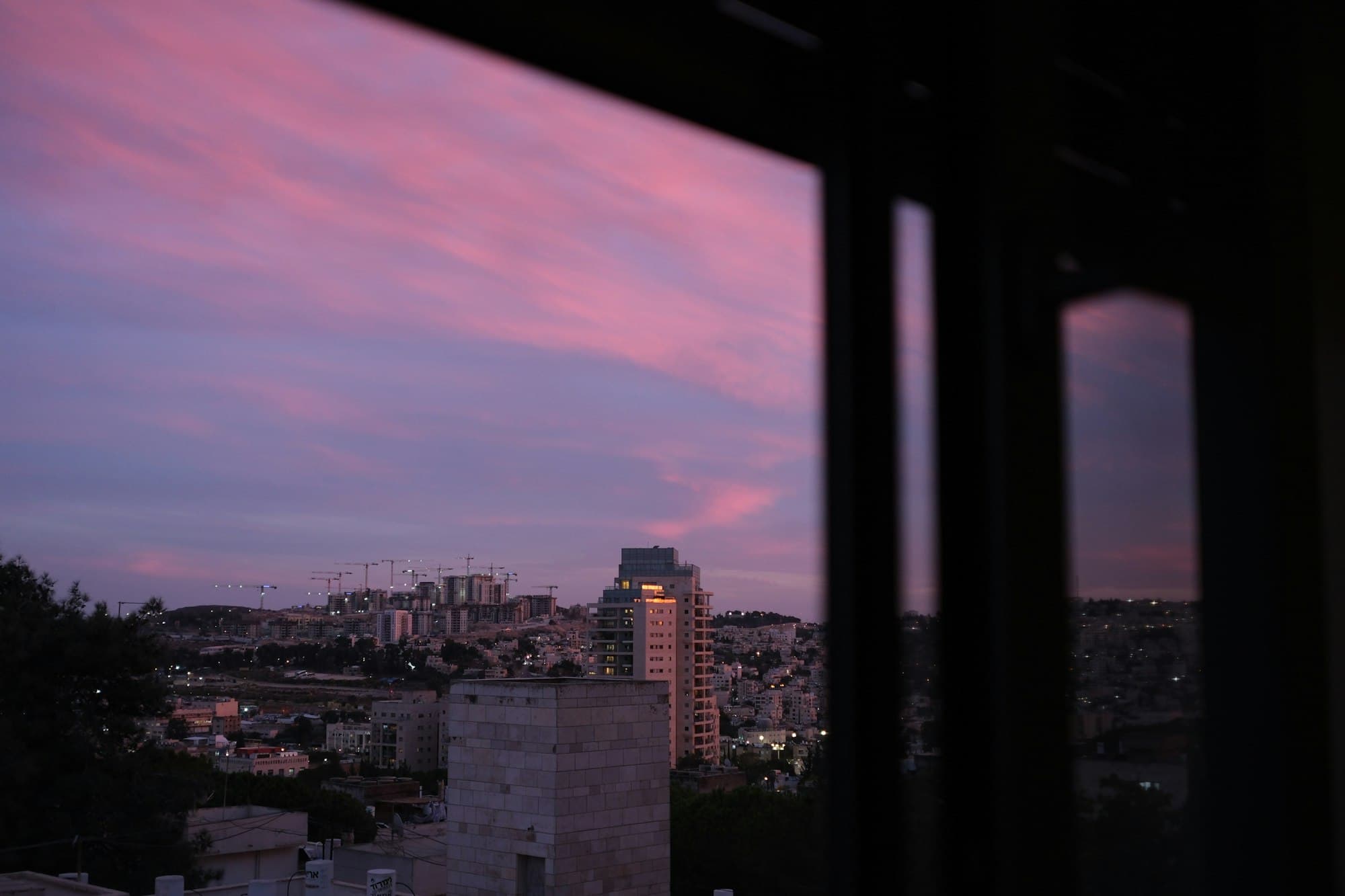 A city skyline with construction cranes under a purple and pink dusk sky seen through a dark window frame