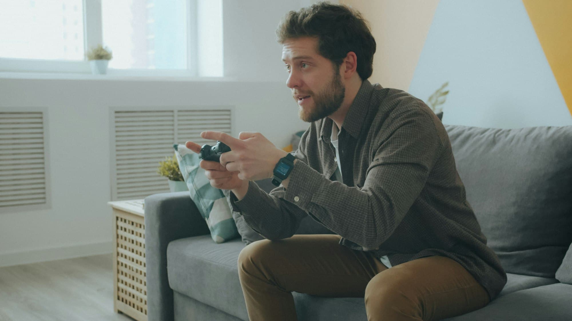 A bearded man on a grey sofa leaning forward and playing a video game with a controller