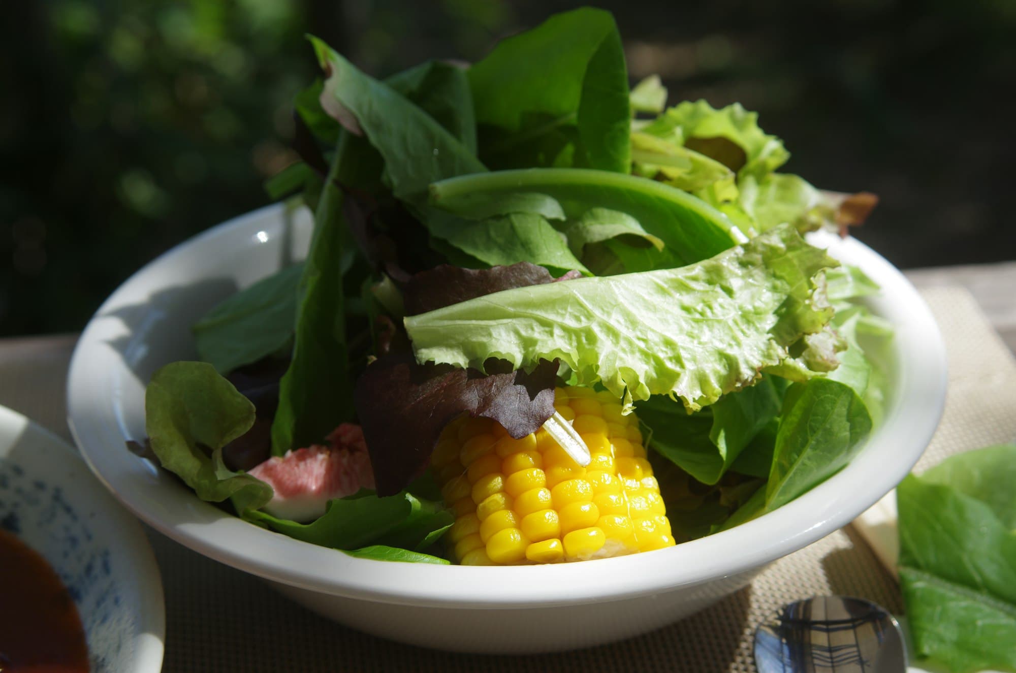 A white bowl of mixed salad greens with sweet corn on a sunlit outdoor table