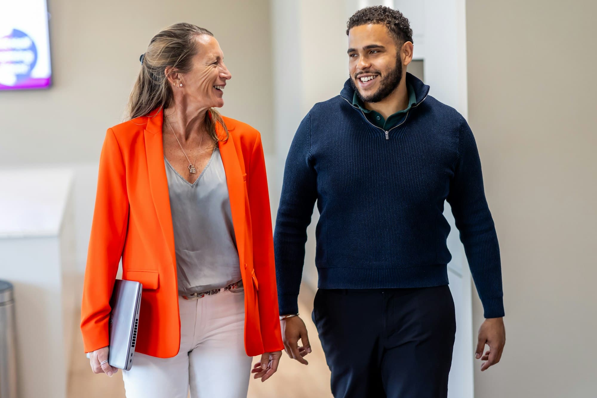 A woman in an orange blazer and a man in a navy sweater walk and smile together in an office