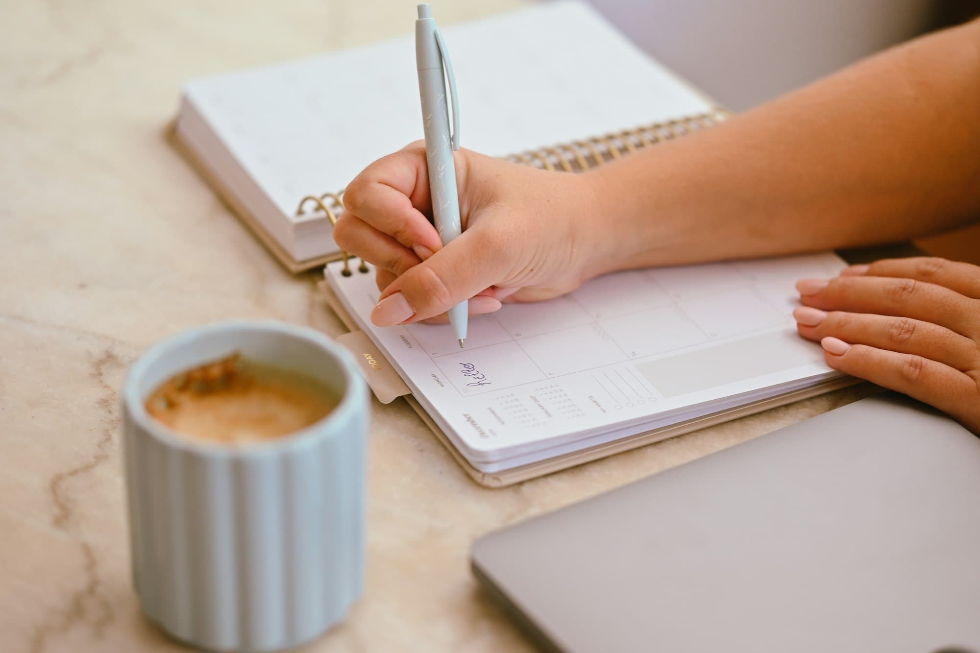 A person writing in a planner with a light blue pen beside a coffee mug